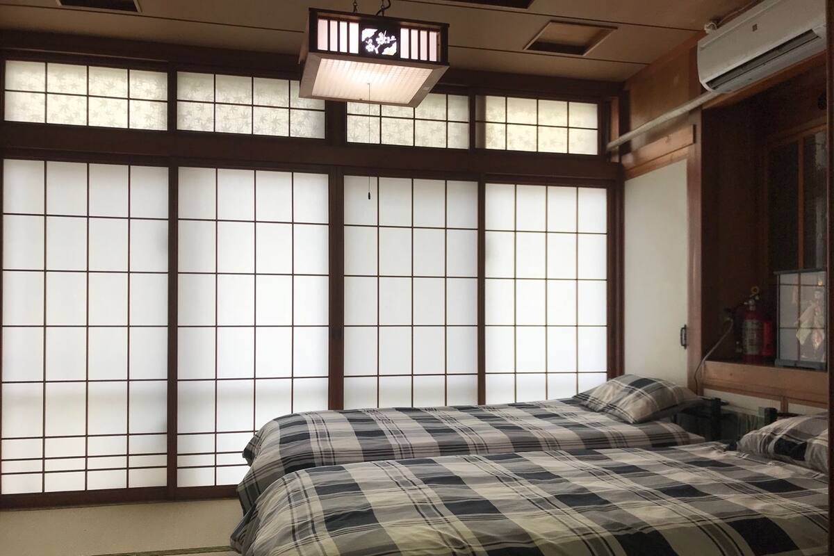 A traditional Japanese room with tatami flooring, a bed with a plaid bedspread, shoji screens, and a hanging light fixture.