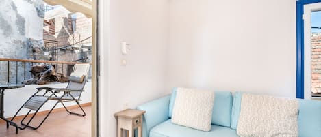 Une pièce avec un canapé bleu clair, deux coussins à motifs, une petite table d’appoint en bois et une vue sur un balcon doté d’un mur en pierre rustique et d’une rambarde en métal.