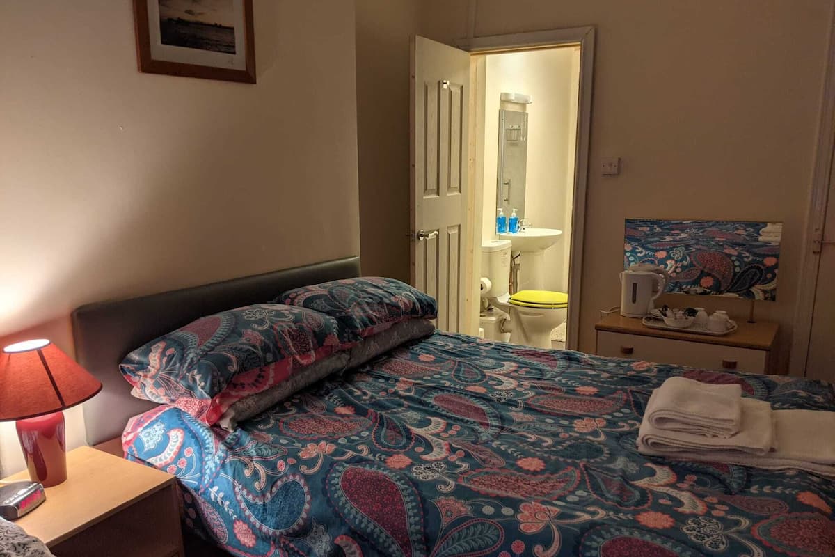 A bedroom with a bed, a nightstand, a lamp, a bathroom visible through an open door, and a small table with a tray of toiletries.