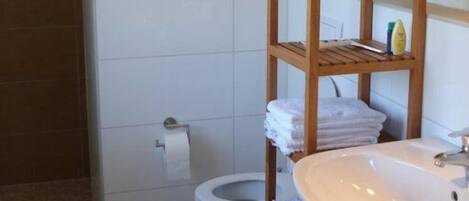 A bathroom with a wooden shelf holding towels and toiletries, a white sink, and a toilet.