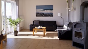 A living room with a sofa, a coffee table, a potted plant, a fireplace, and a large window. - Strandleben Fehmarn - Ferienhaus 3 (Fehmarn)
