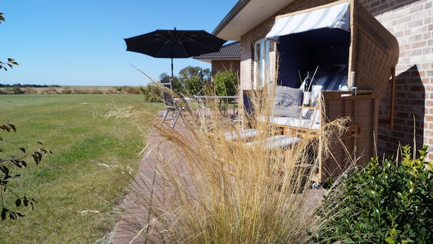 A patio area with a wicker chair and an umbrella, a brick house, and a grassy field in the background. - Strandleben Fehmarn - Ferienhaus 3 (Fehmarn)