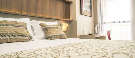 A hotel room with a large bed, wooden headboard, and a view of the city through sheer curtains.