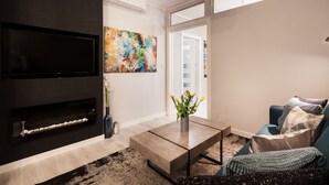 Una sala de estar moderna con un televisor de pantalla plana, un cuadro abstracto de colores, un florero con flores y un sofá azul con cojines.
