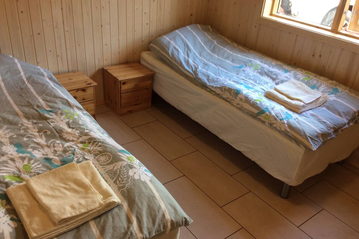 A small, cozy room with two single beds, each with a wooden nightstand. The room has wooden walls and a window providing natural light.