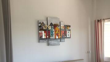 Un salon avec un canapé beige, une table basse et une œuvre d’art fixée au mur représentant des maisons aux couleurs vives.