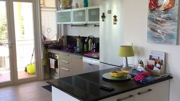 Une cuisine moderne dotée d’un îlot central, de meubles blancs, d’un four à micro-ondes et d’un réfrigérateur. Une fenêtre offre une vue sur l’extérieur, et un tableau est accroché au mur.