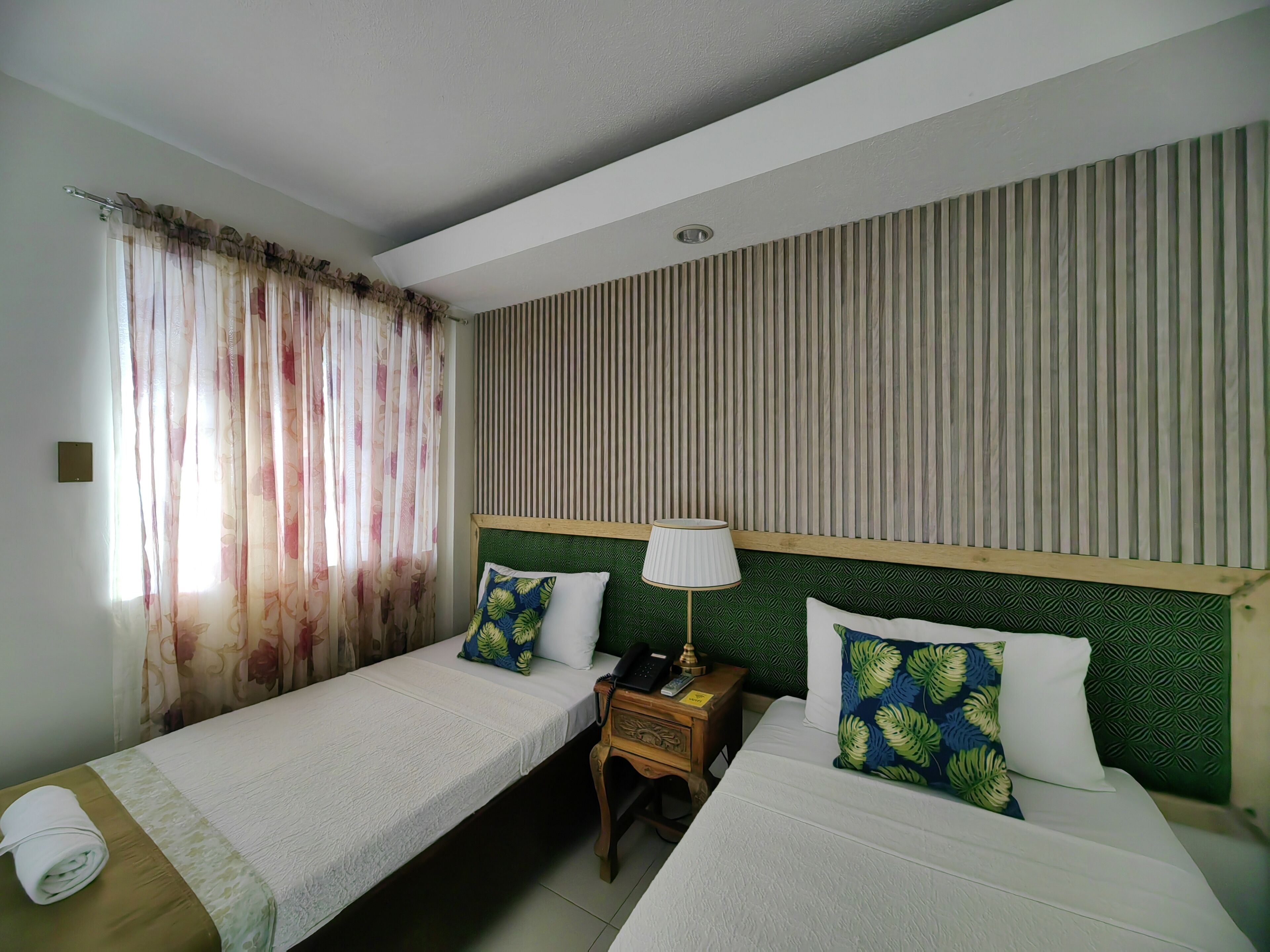 Habitación de hotel con dos camas individuales, una mesita de noche de madera con lámpara, una pequeña mesa con una toalla y una ventana con cortinas de encaje.