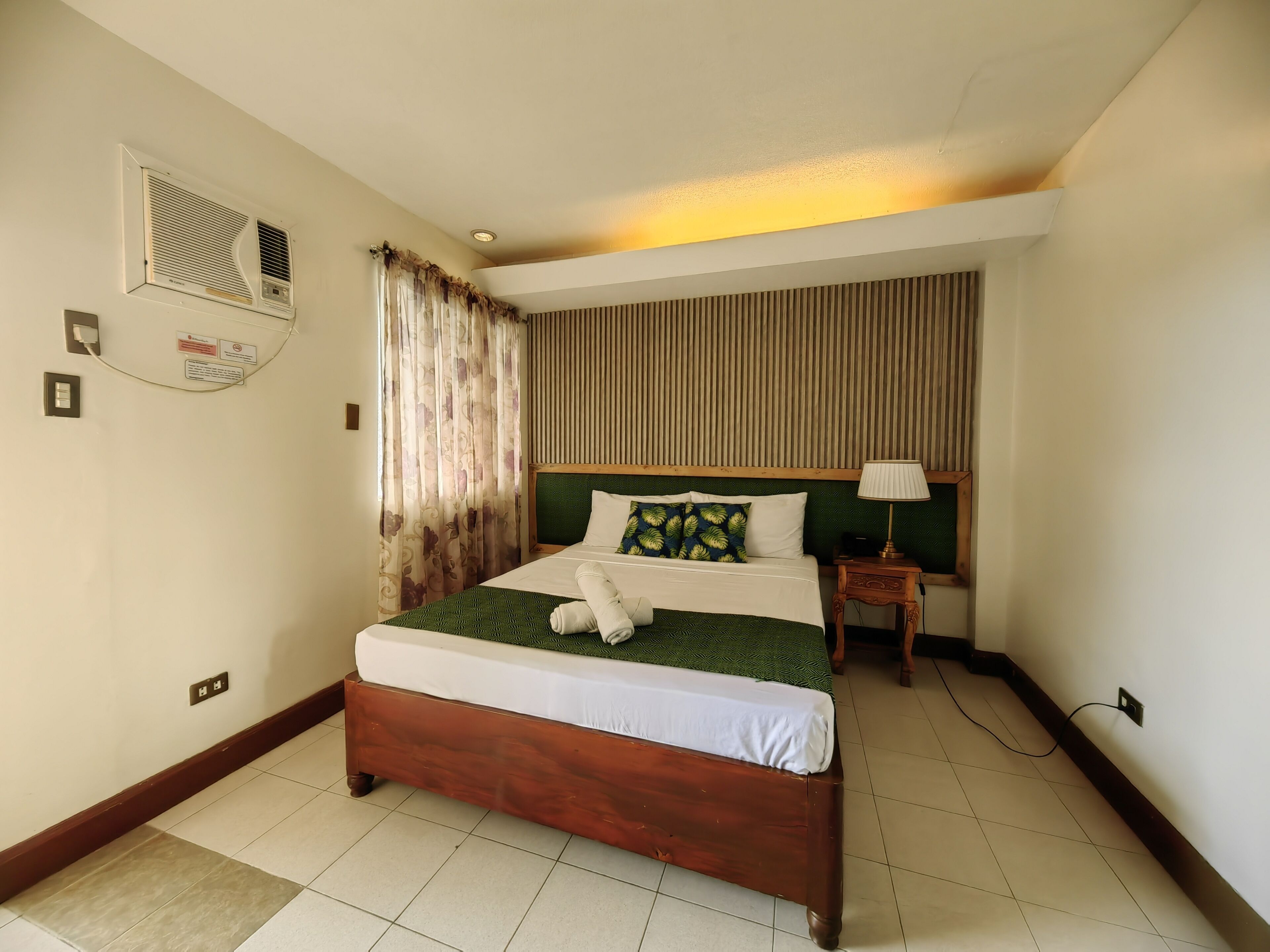 Ein Hotelzimmer mit einem hölzernen Bett, einem Nachttisch, einer Lampe, einem Fenster mit Vorhängen und einer Wand-Klimaanlage.