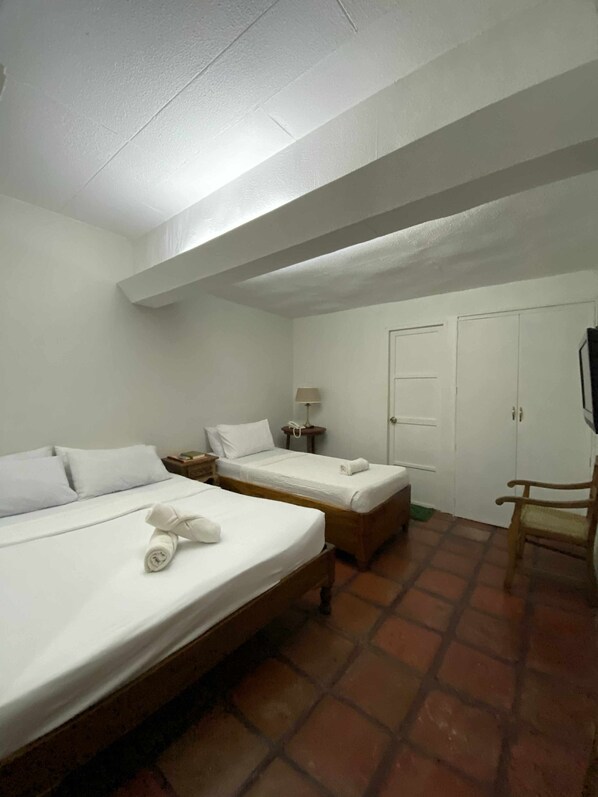 Habitación de hotel con dos camas, una silla de madera, una mesita de noche con lámpara y un armario.