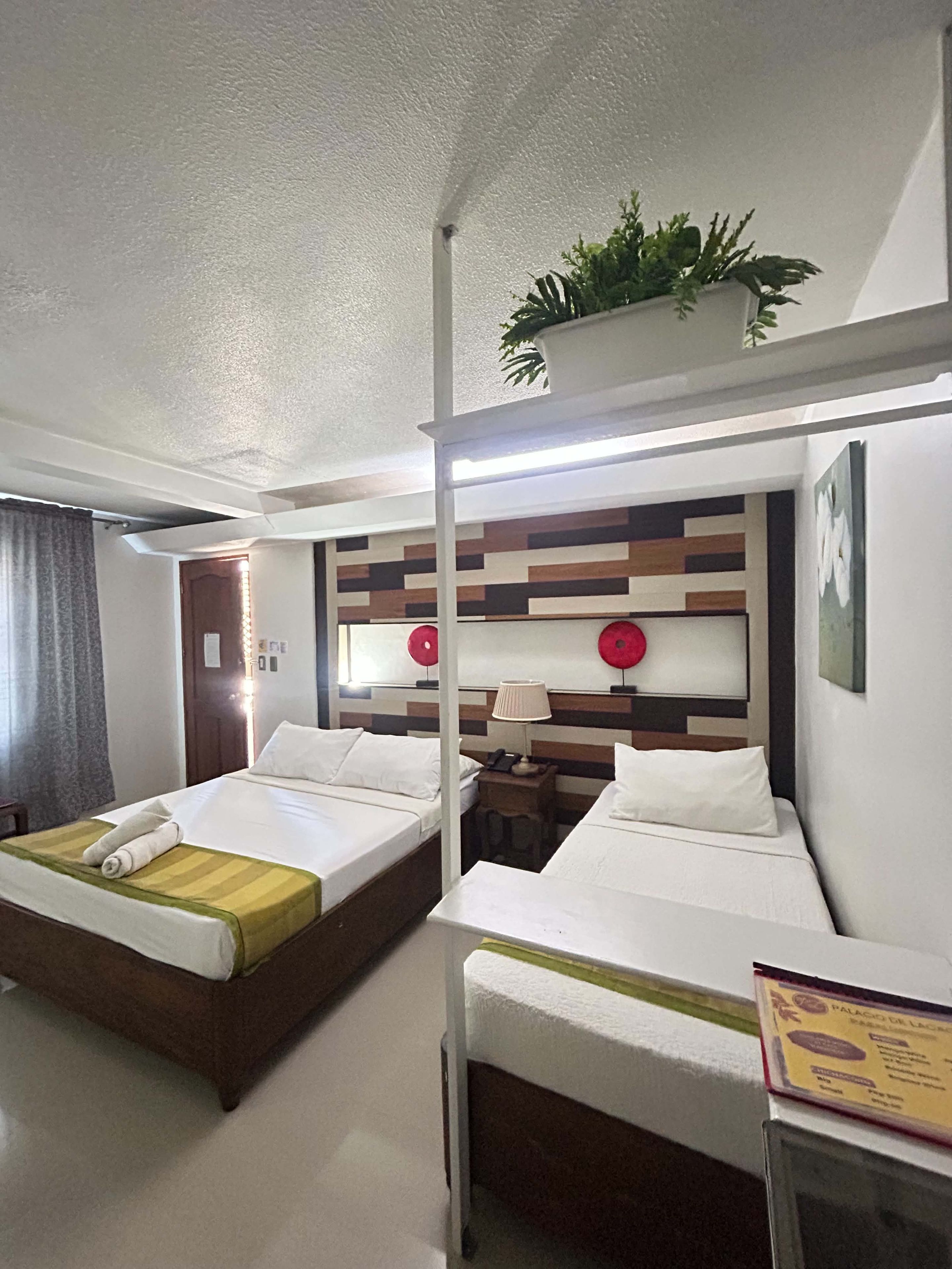 Habitación de hotel con dos camas, una mesita de noche con lámpara, una pequeña mesa con un menú y una planta en maceta en la pared.