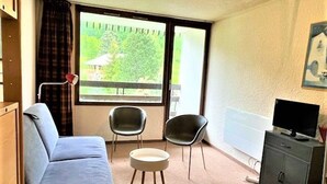 Un espace de vie compact comprenant un canapé, deux fauteuils, une petite table ronde, un téléviseur posé sur un meuble et une fenêtre donnant sur un paysage verdoyant.