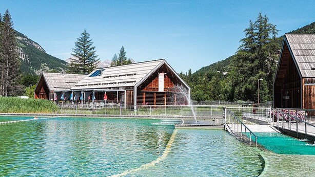 Une piscine avec un bâtiment en bois en arrière-plan, entourée d’arbres et de montagnes.