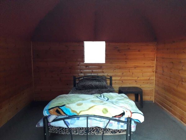 A small, cozy room with wooden walls and a sloped ceiling. It features a single bed with a patterned blanket and a small bedside table.