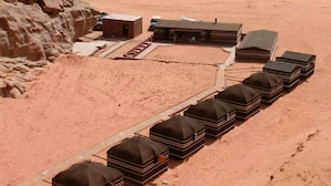 A desert landscape with a row of black tents aligned along a pathway, a small building, and a vehicle in the background. - Desert Lovers Camp (Wadi Rum)