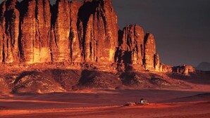 A desert landscape with a prominent rock formation in the background and a person standing in the foreground. - Desert Lovers Camp (Wadi Rum)