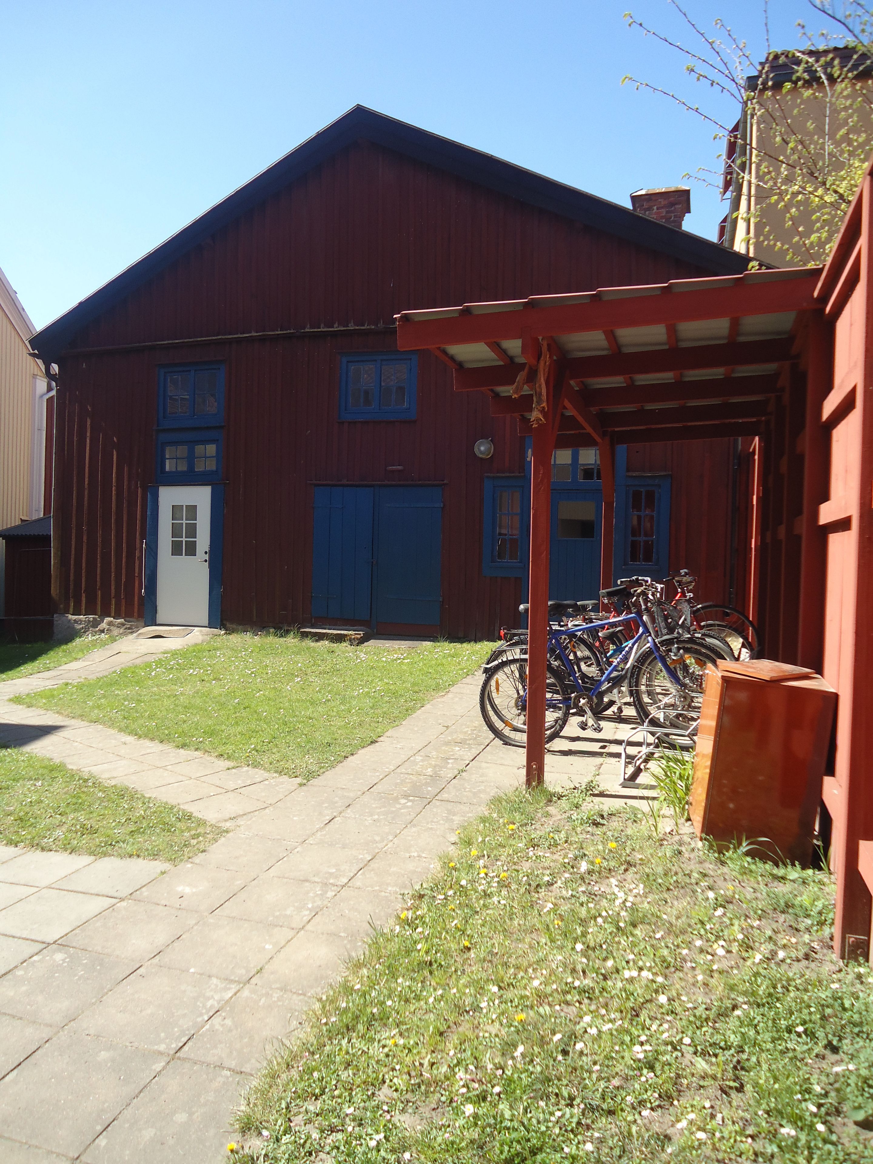 A red wooden building with a covered porch, a white door, and a blue garage door. There are bicycles parked outside.
