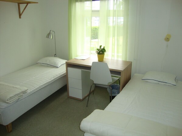 A dormitory room with two single beds, a desk with a chair, a lamp, and a potted plant.