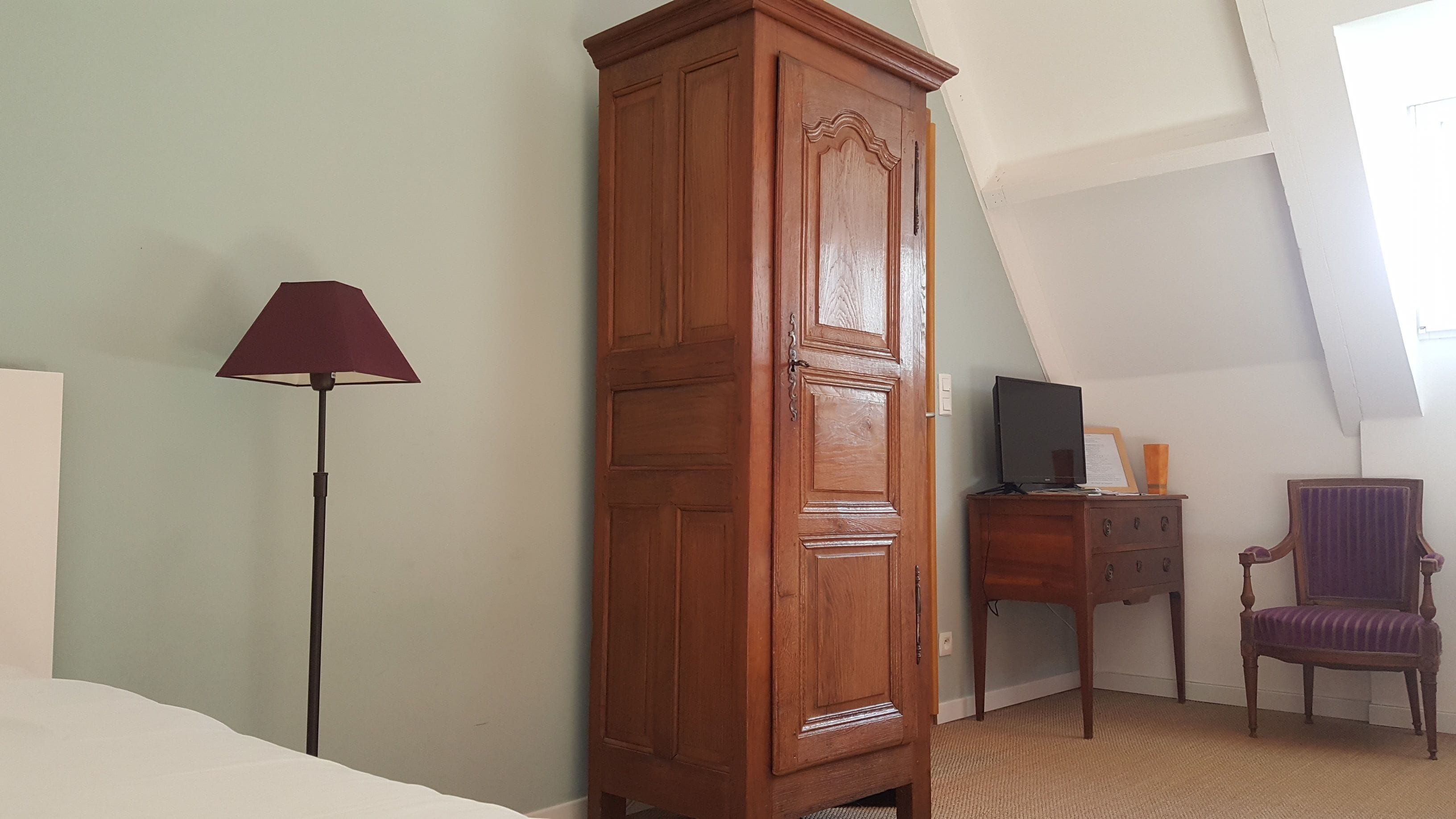 Ein Zimmer mit einem hohen Holzschrank, einem Schreibtisch mit Computer, einem Stuhl und einer Stehlampe.