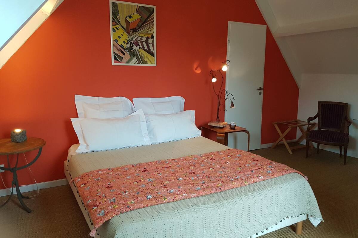A bedroom with a bed, a chair, a small table, a wall painting, and a door.
