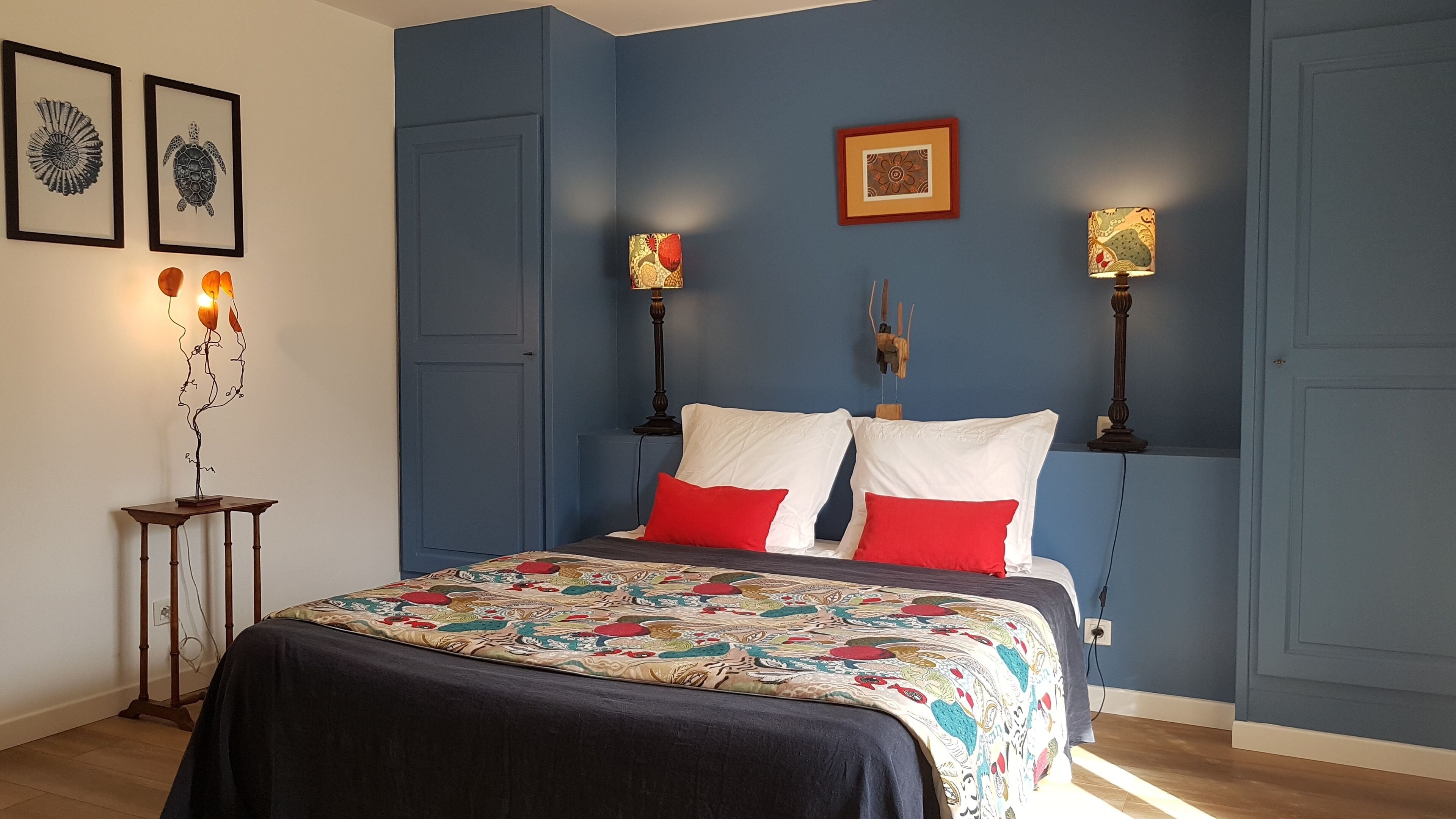 A bedroom with a bed, two bedside lamps, a wall-mounted clock, and framed artwork.