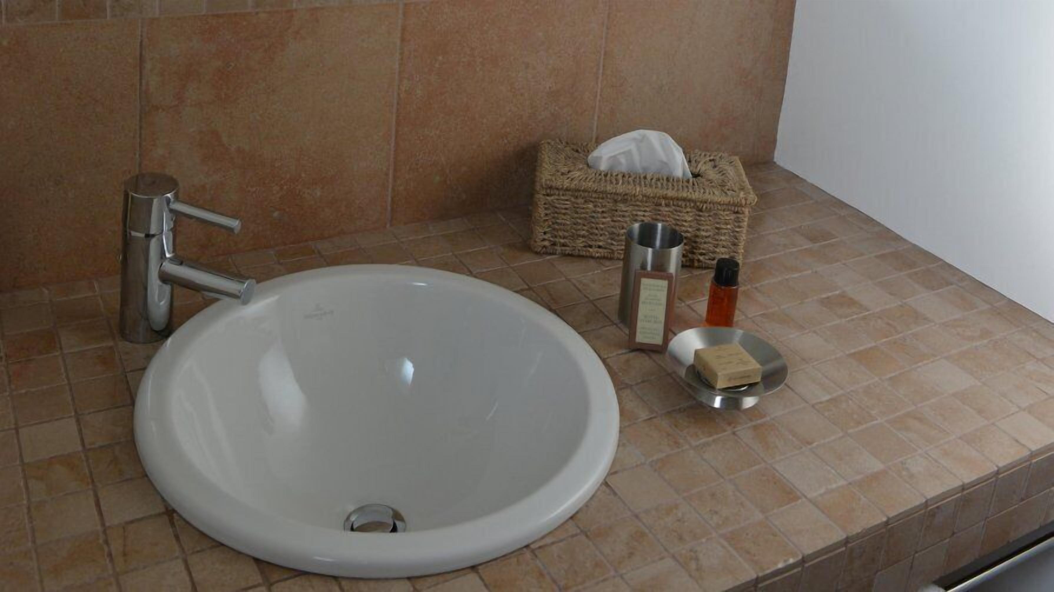 Un baño con un lavamanos redondo blanco, un grifo plateado, una caja de pañuelos, un jabonero y una botella pequeña.