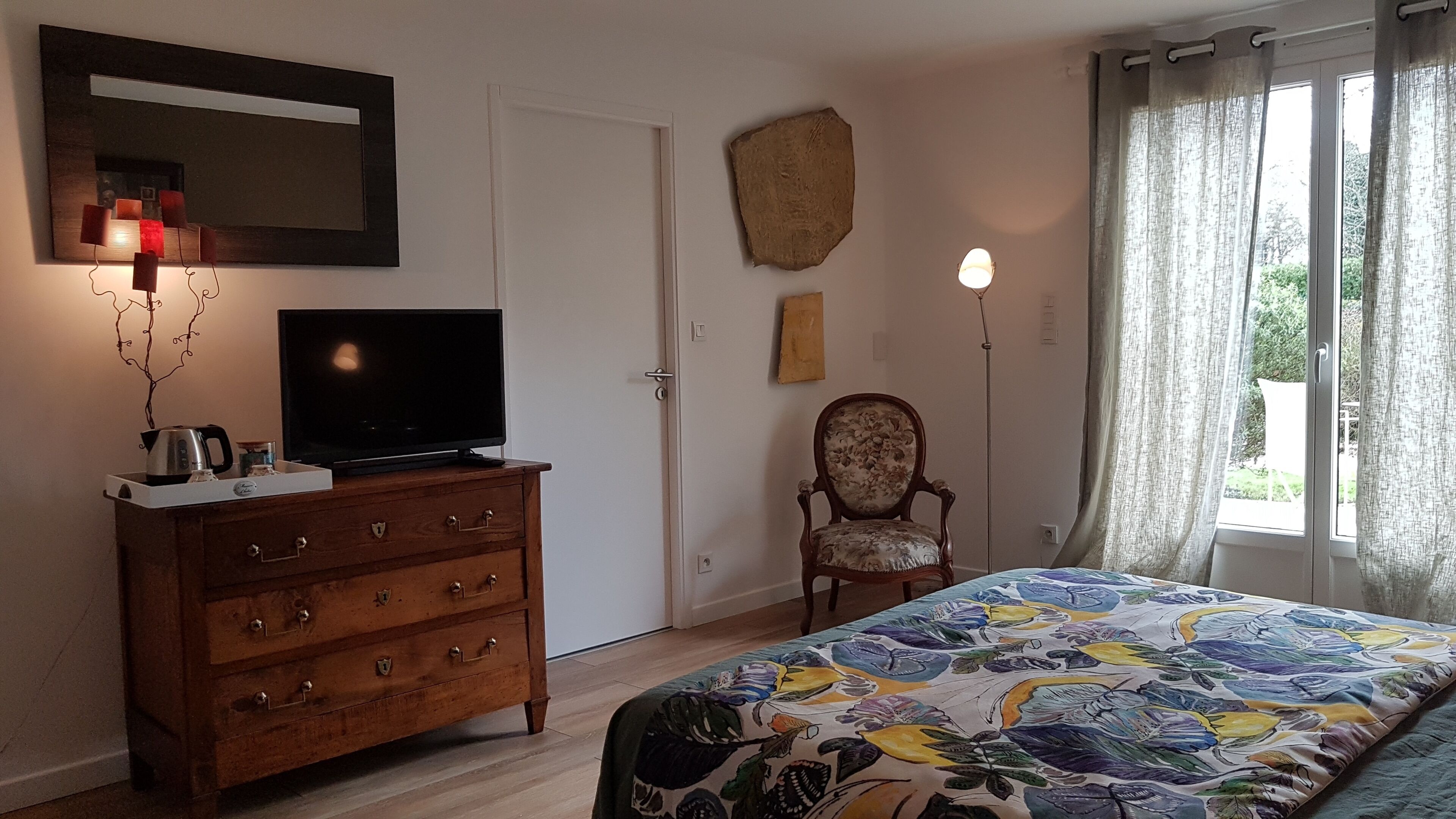 Une chambre à coucher avec une commode en bois, un téléviseur à écran plat, une couvre-lit à motifs floraux, une chaise et une grande fenêtre avec des rideaux.