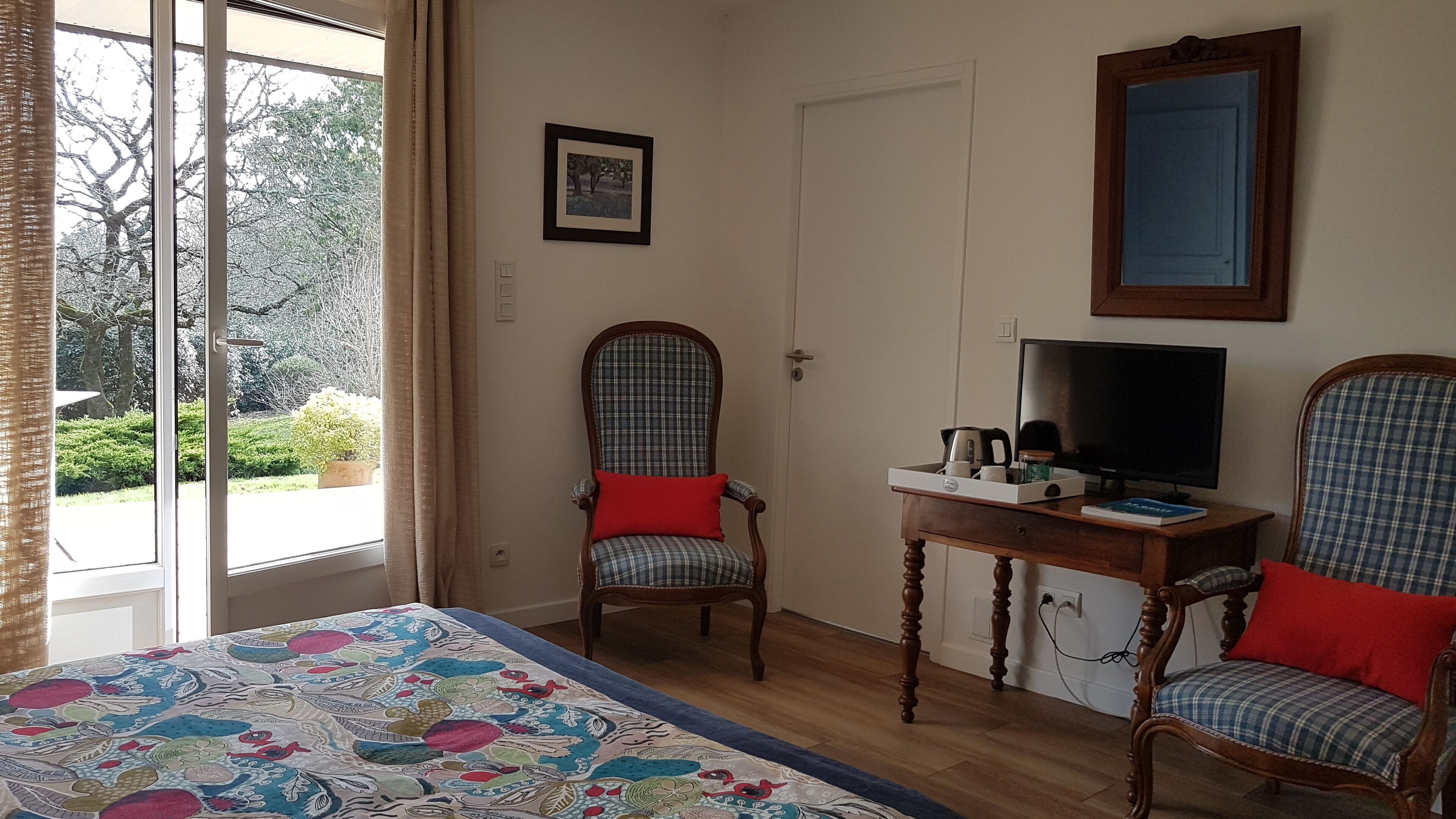 Habitació amb un llit, dues cadires, una taula amb televisió i un plat de coses, un quadre a la paret, i una finestra amb cortines.