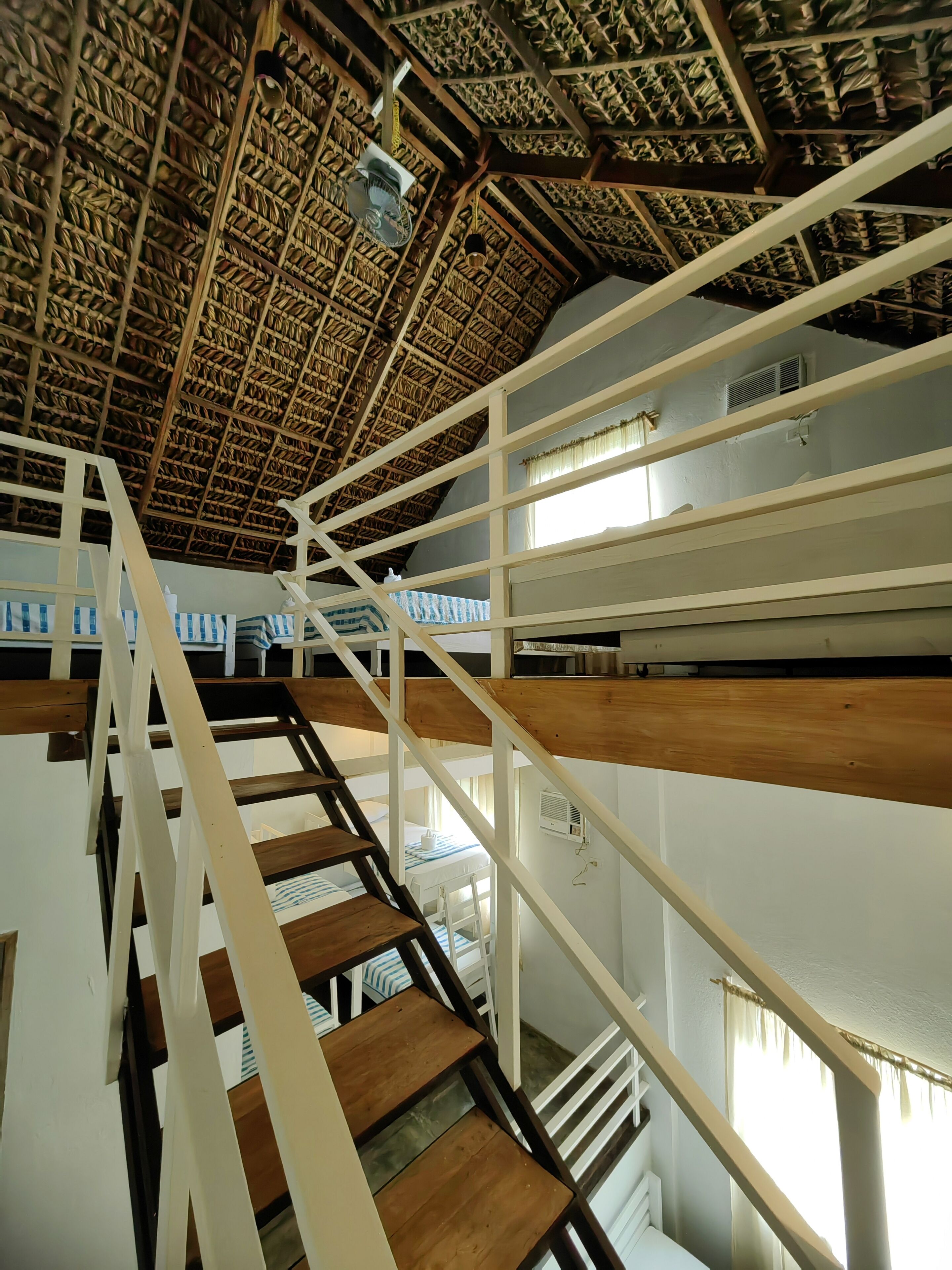 A staircase with wooden steps and white railings leading to a room with a thatched roof ceiling and a bed with a striped blanket.