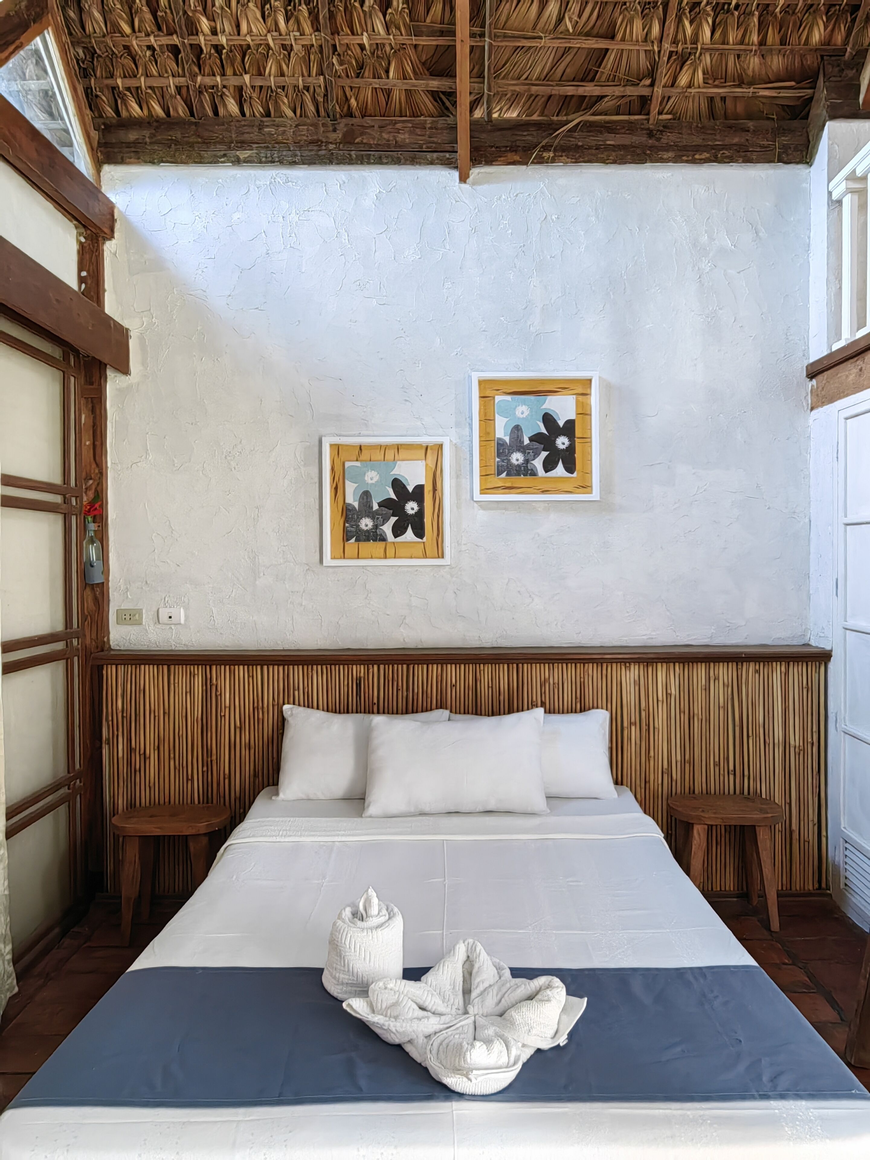 A bedroom with a bed, wooden headboard, two wall-mounted flower paintings, and a window with a wooden frame.