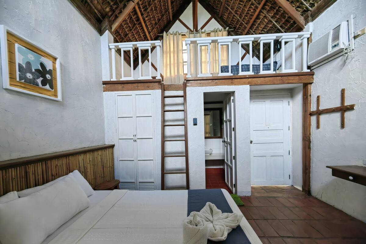 Une chambre à coucher avec un lit, une tête de lit en bois, un balcon avec une balustrade et une vue sur l’extérieur.