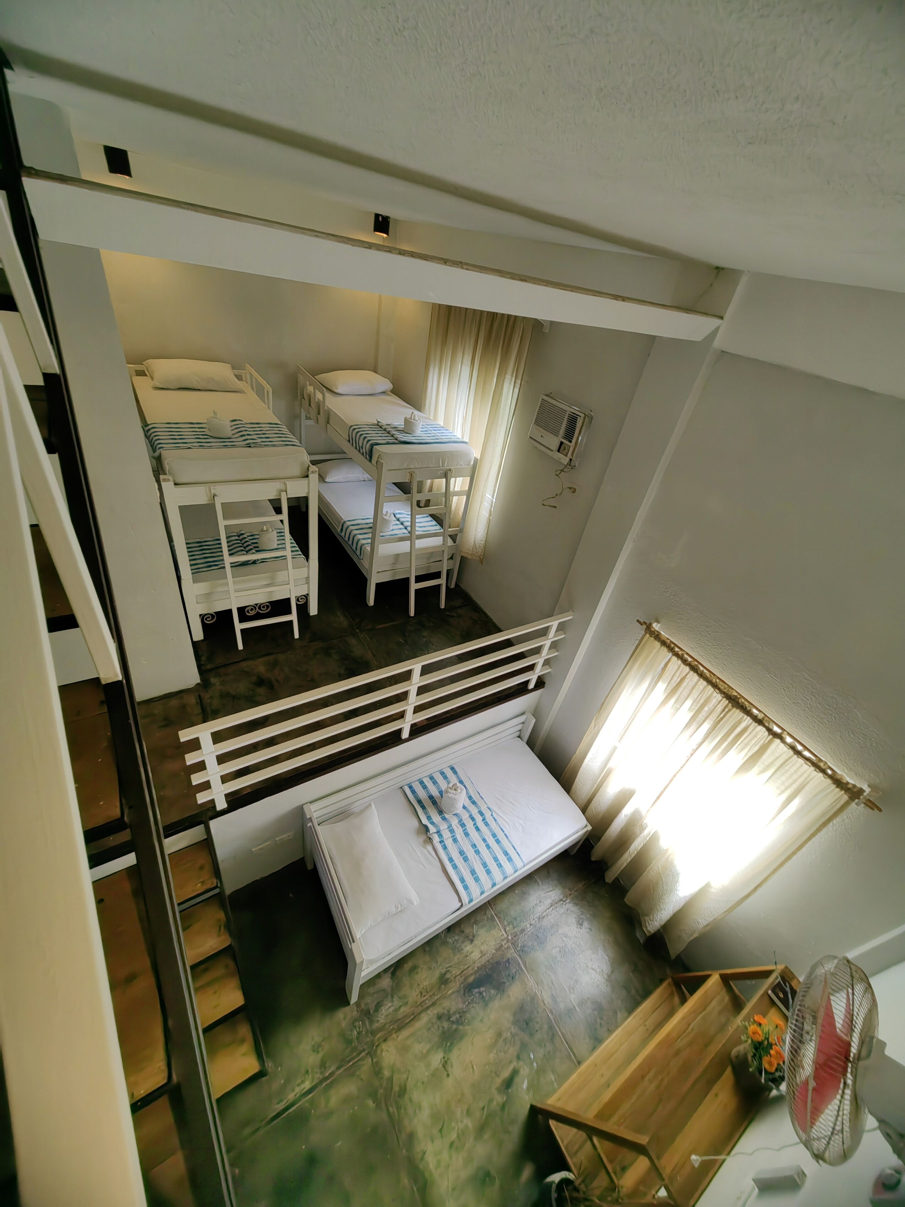 A dormitory room with bunk beds, a small table with a fan, and a window with curtains.