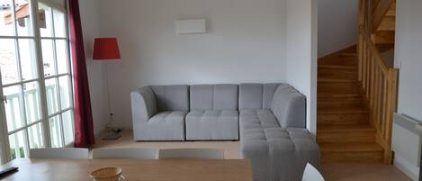 A modern living room with a grey sectional sofa, a wooden dining table, and a staircase.
