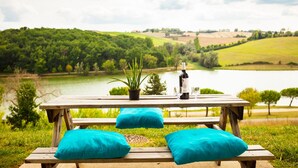 A wooden picnic table with turquoise cushions, a potted plant, and a bottle of water, overlooking a lake and green hills.