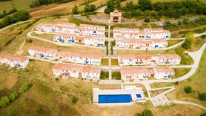 An aerial view of a residential area with multiple white houses with red-tiled roofs, arranged in a semi-circular pattern around a central courtyard with a swimming pool.
