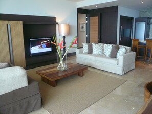 A modern living room with a flat-screen TV, a coffee table with a vase of flowers, a sofa with patterned cushions, and a kitchen area in the background.
