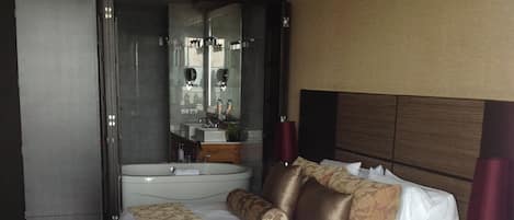 A hotel room with a large bed, a sitting area, a bathroom with a mirror and sink, and a view of the hallway.