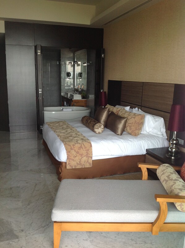 A hotel room with a large bed, a sitting area, a bathroom with a mirror and sink, and a view of the hallway.
