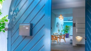 Uma porta azul com caixa de correio, uma sala de jantar com cadeiras de vime e uma mesa, e um lustre.