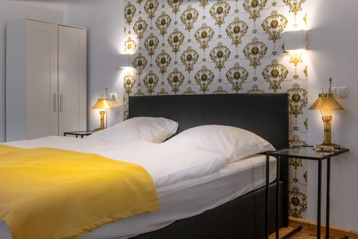 A neatly made bed with a yellow blanket, a bedside table with a lamp, a wardrobe, and a bathroom with a sink and mirror.