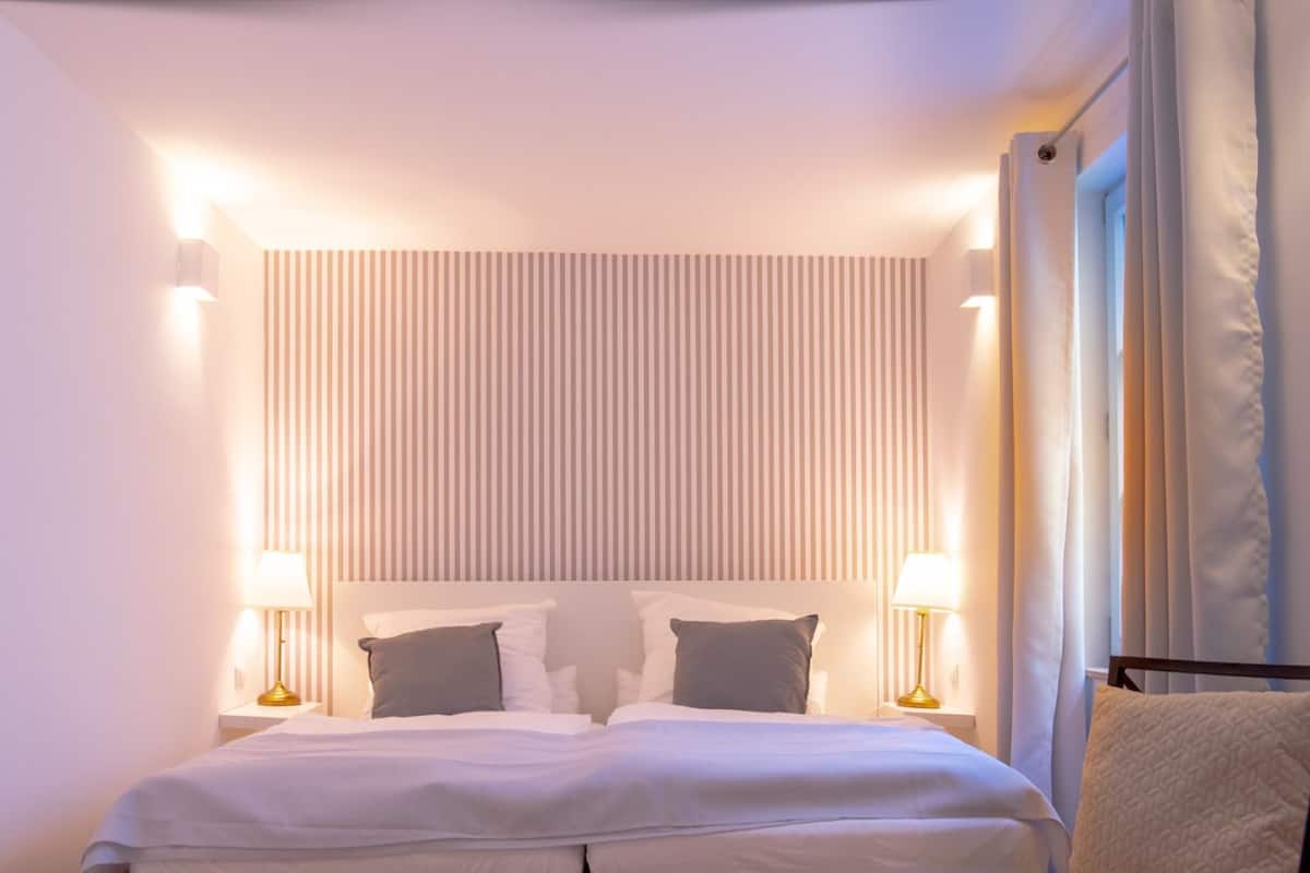 A hotel room with a bed, two bedside lamps, a headboard with vertical stripes, and a window with curtains.
