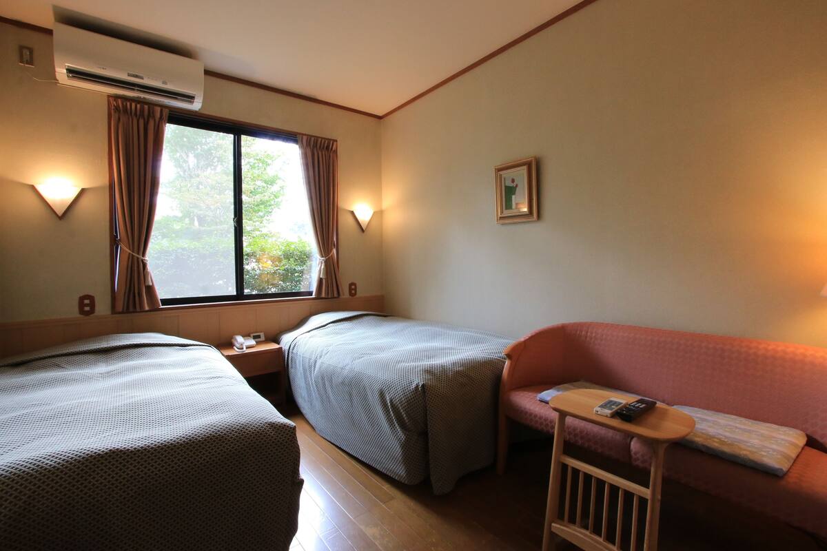 A hotel room with two single beds, a sofa, a small table with a remote control, and an air conditioning unit.