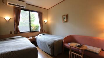 A hotel room with two single beds, a sofa, a small table with a remote control, and an air conditioning unit.