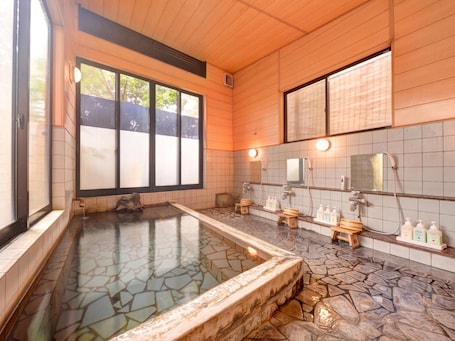 A traditional Japanese onsen with a stone hot spring, wooden benches, and a tiled wall with mirrors and toiletries.