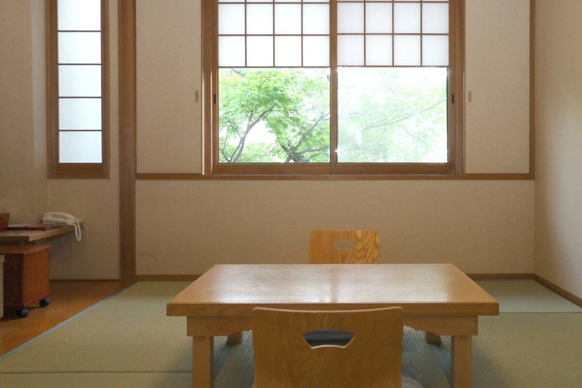 A room with tatami flooring, a wooden table, a chair, and a window with a view of trees.
