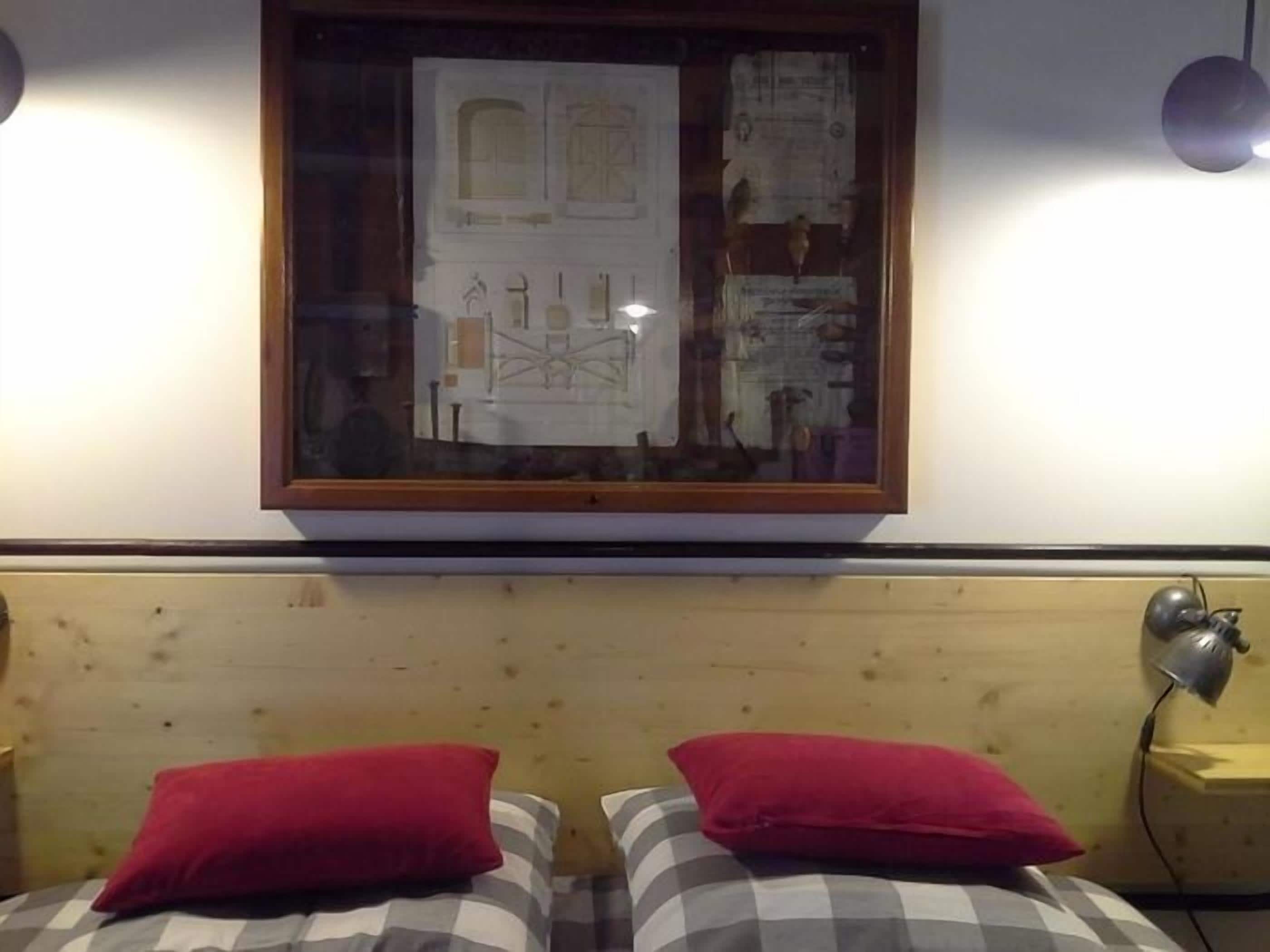 A room with a bed featuring a checkered bedspread and two red pillows. Above the bed, there is a framed picture on the wall.