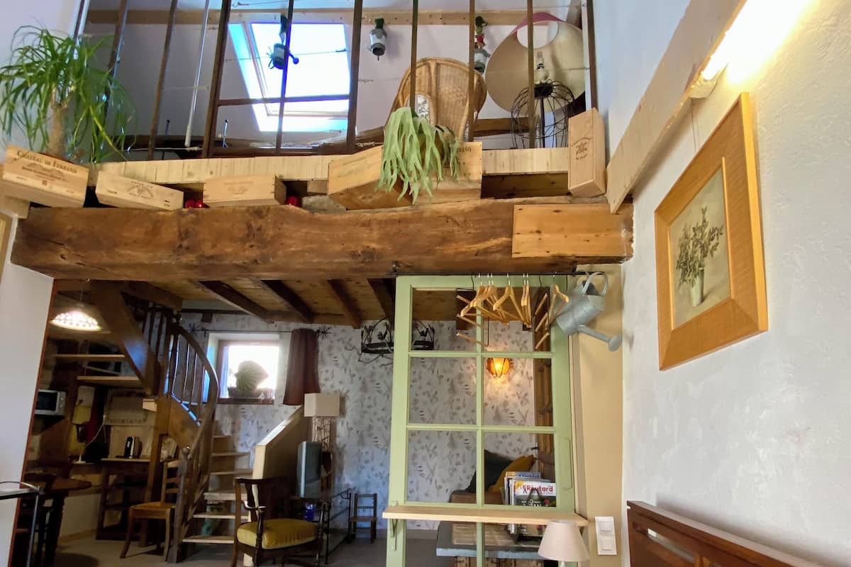 Une chambre de style « loft » avec un plafond en bois, un lit recouvert d’une literie à motifs floraux, un fauteuil vert et un escalier menant à une mezzanine.