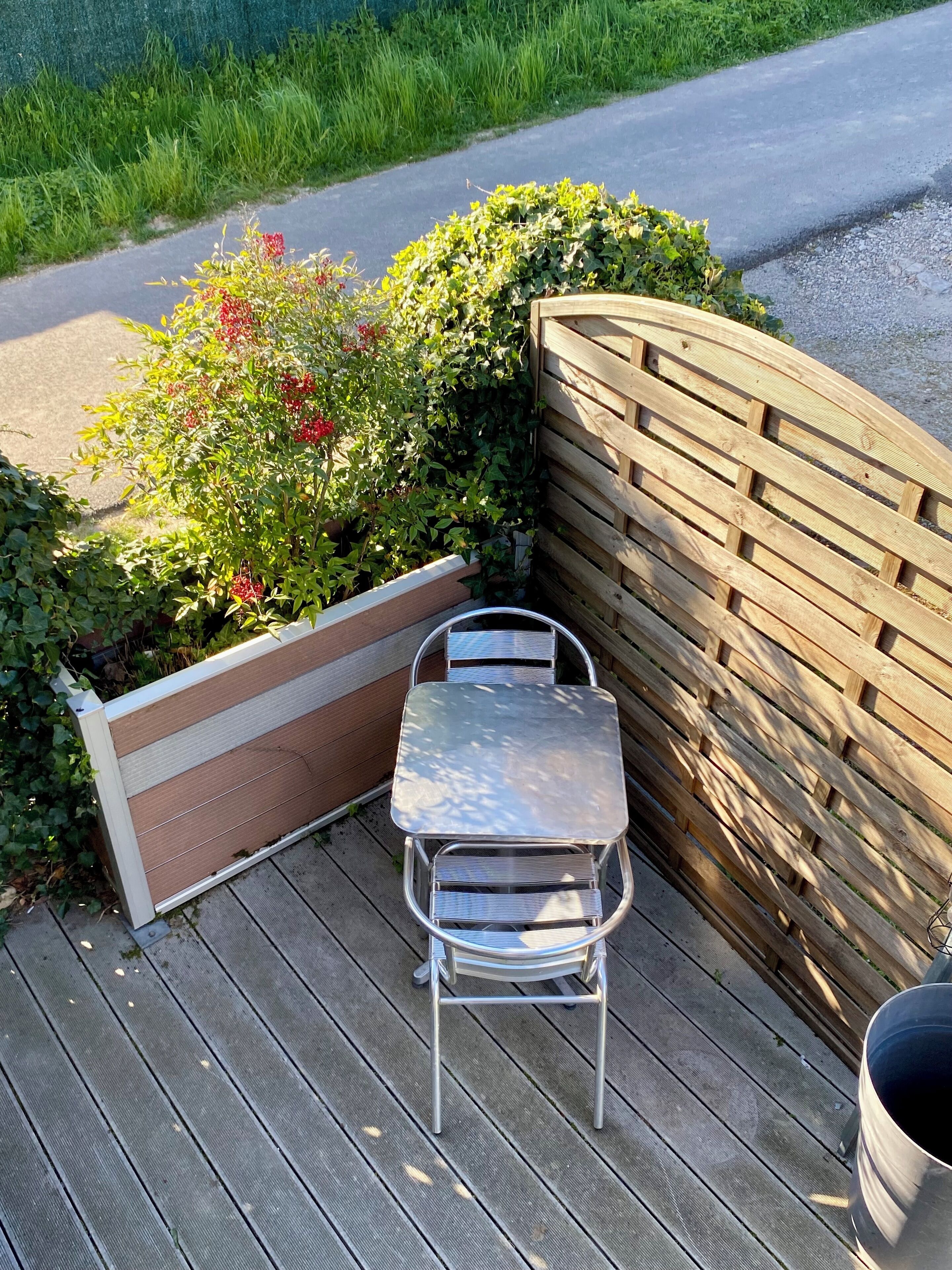 A corner outdoor seating area with a metal table and chair, surrounded by a wooden fence and greenery.