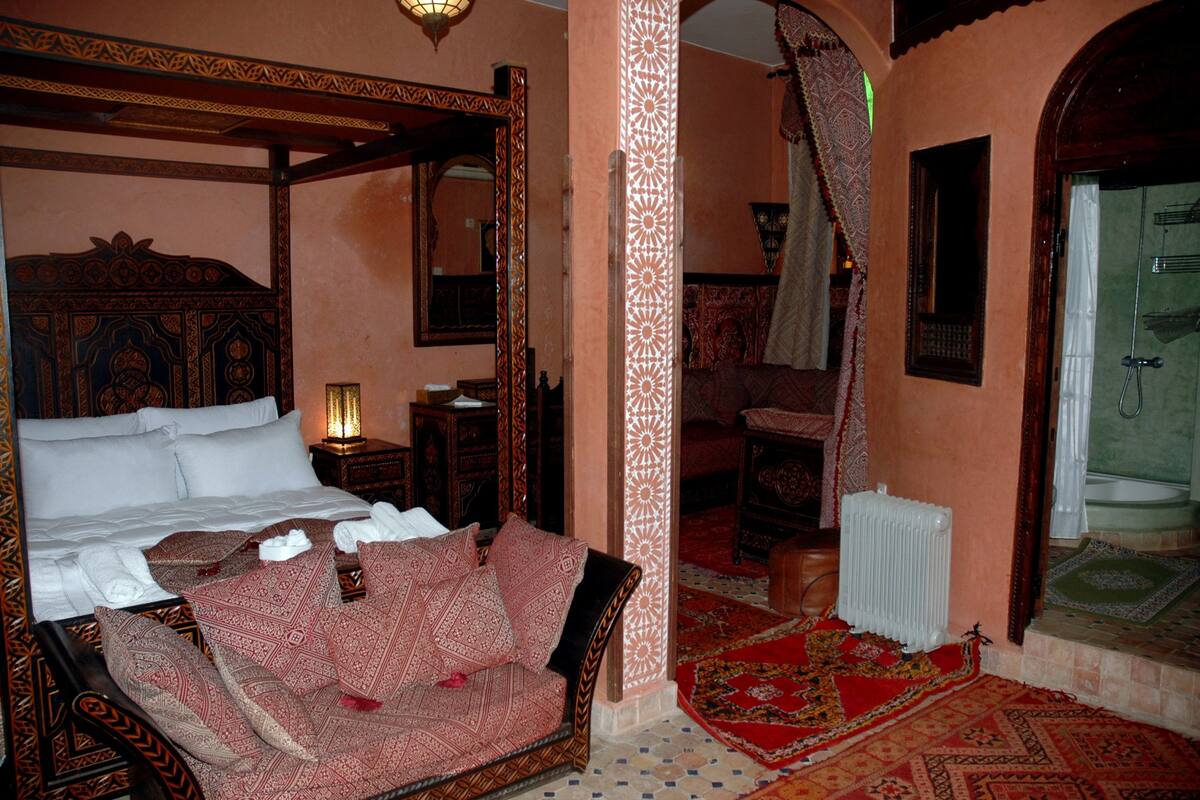 A traditional room with a four-poster bed, a sitting area with a patterned sofa, and a view of a bathroom with a shower and a radiator.