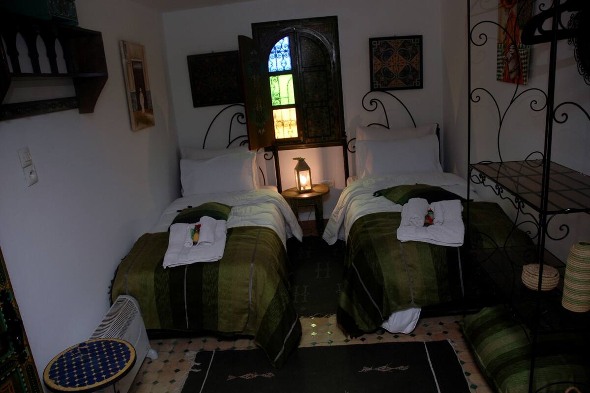 A small, dimly lit room with two beds, a nightstand with a lantern, and a decorative window.